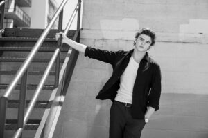 Black and white senior portrait of young man leaning on industrial stair railing in Los Angeles.