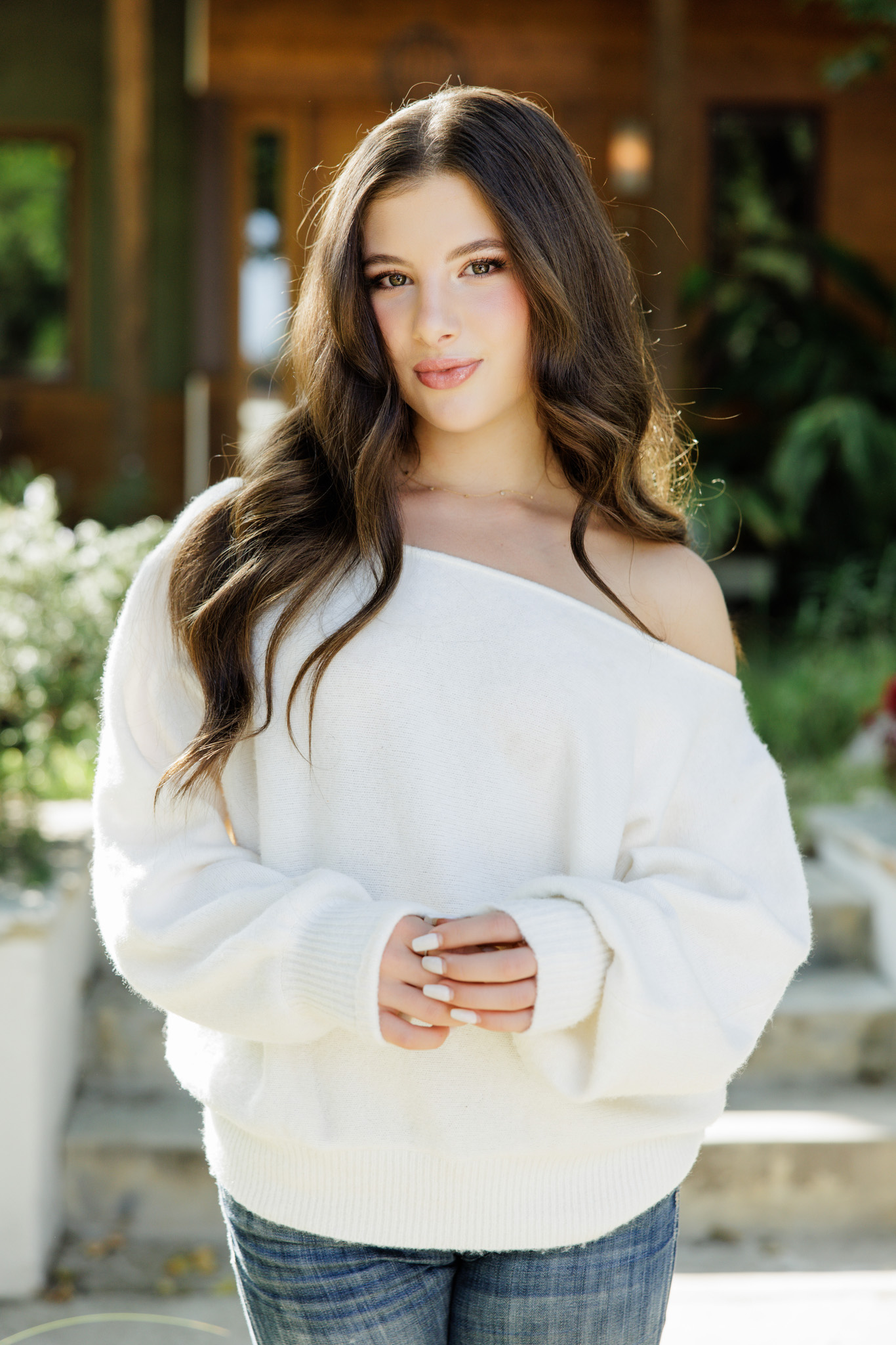 Brunette senior girl in an off-the-shoulder white sweater and jeans sitting on a stone ledge and smiling in front of a craftsman-style home in Studio City during her senior portrait session