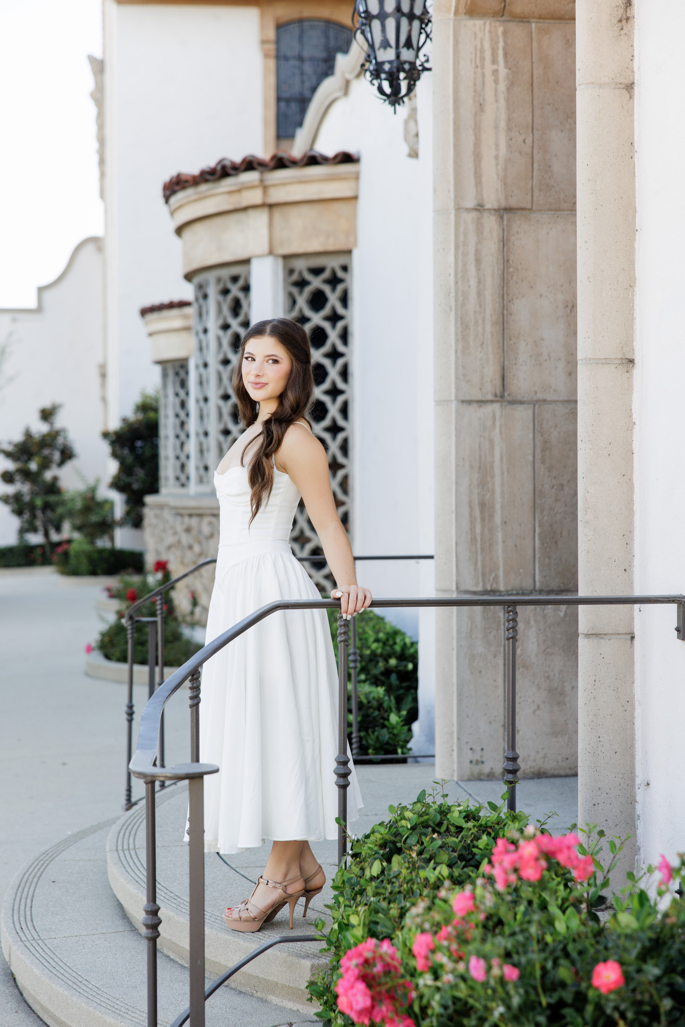 Editorial senior portrait of a girl in a white midi dress and nude heels leaning on an iron railing in front of Spanish-style architecture with ornate lattice windows and pink roses in the San Fernando Valley
