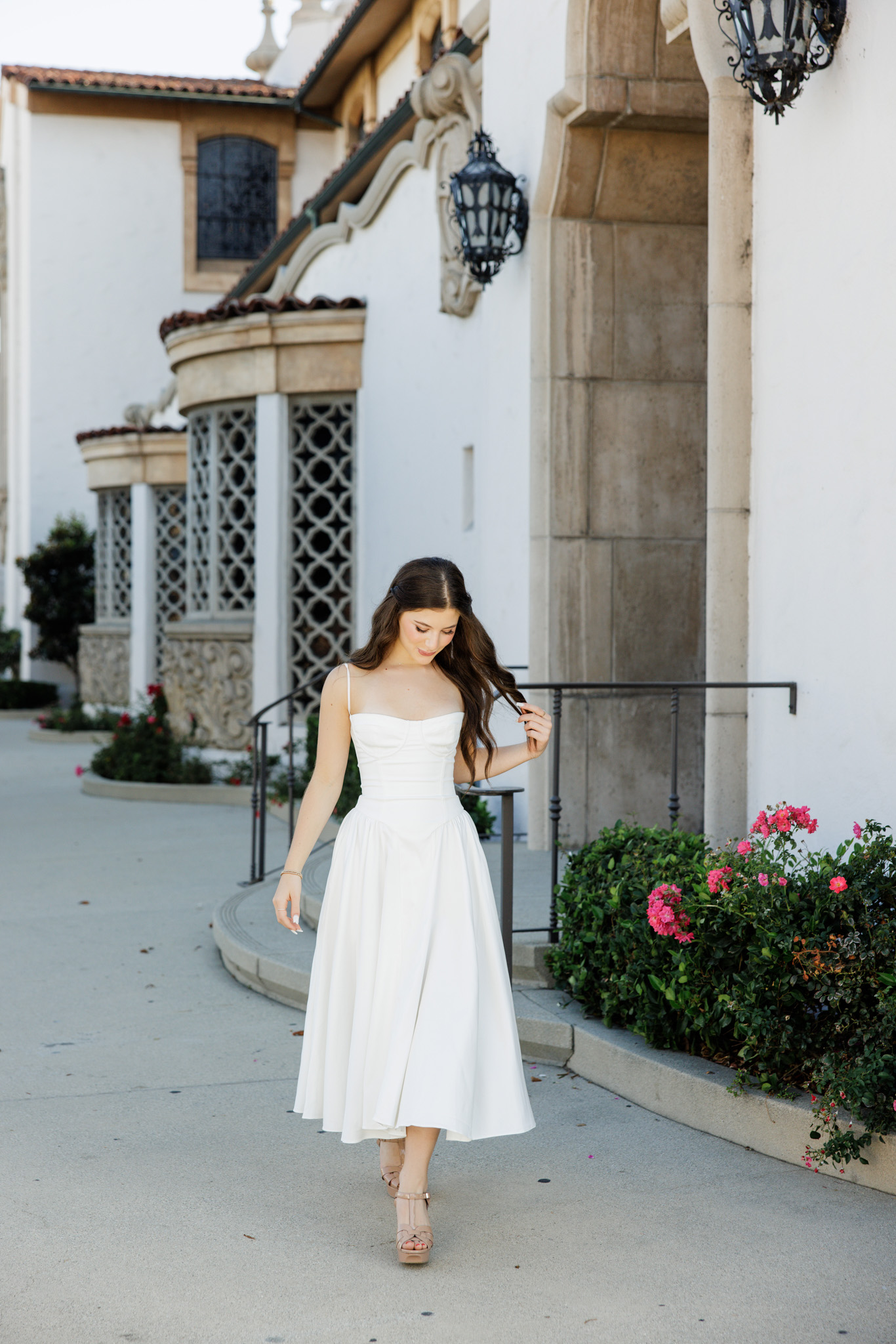Senior girl in a white corset dress walking along a curved sidewalk past stone columns and wrought iron details at a Spanish Revival building during her senior pictures in the San Fernando Valley