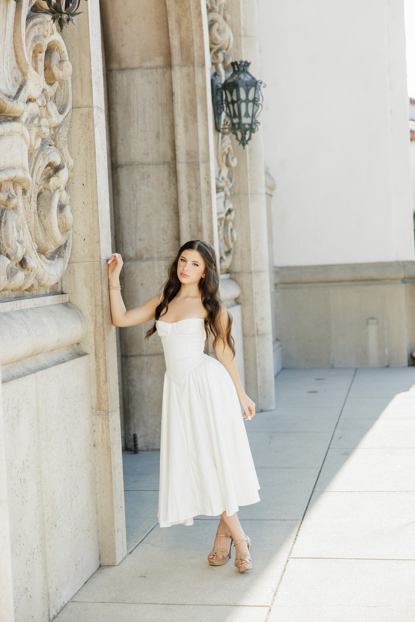 Full-length senior portrait of a girl in a white midi dress and platform heels posing against carved stone architecture with afternoon light streaming through during her San Fernando Valley senior session