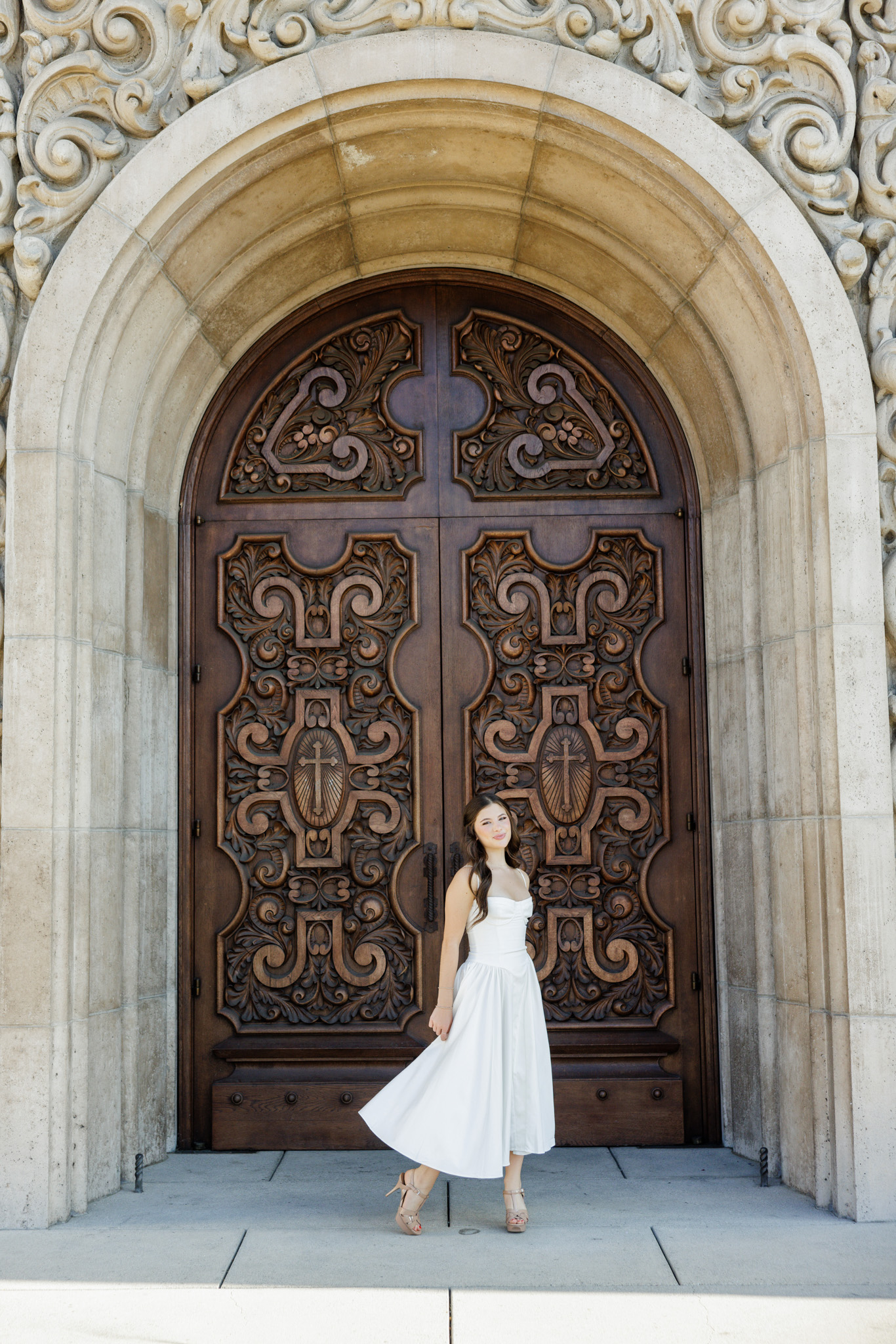 Senior girl in a white dress walking in front of massive carved wooden doors under an ornate stone archway during her on-location senior photo session in the San Fernando Valley