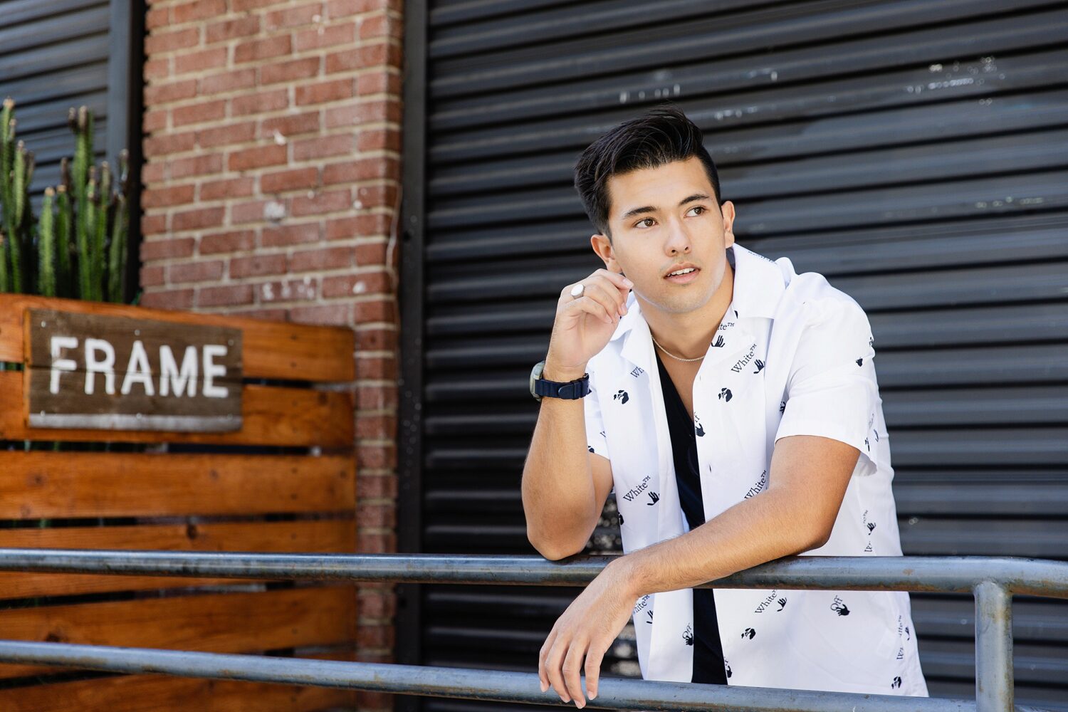 Senior guy leaning on a metal railing in Downtown Los Angeles wearing a white printed button-up and black tee