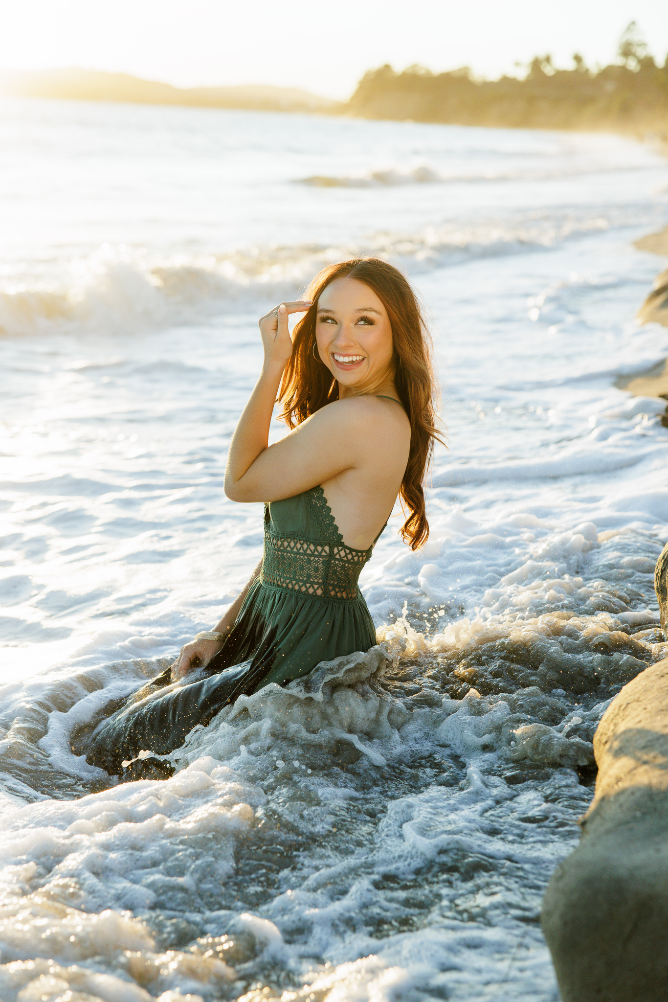Senior laughing while sitting in the ocean waves in a green lace-detail dress with rocks and golden sunset light behind her