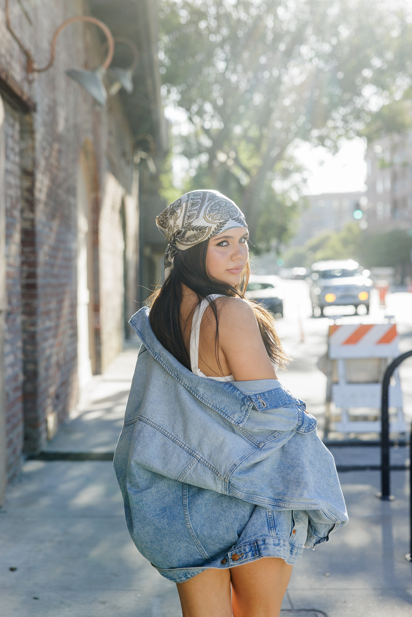 teen girl looking back over her shoulder on a Pasadena sidewalk with her denim jacket draped low and a printed bandana headscarf