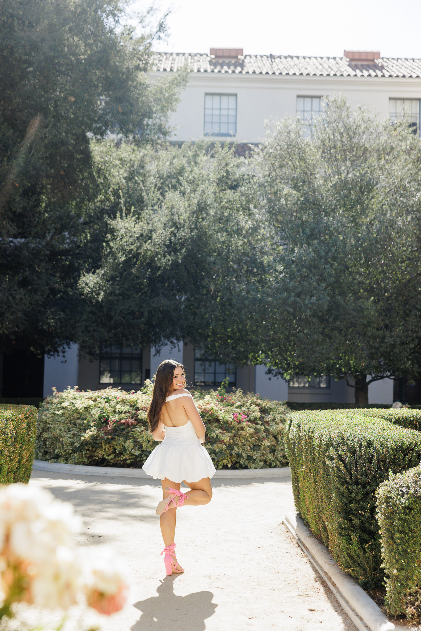 teen senior girl looking back over her shoulder in a white off-the-shoulder mini dress and pink lace-up heels walking down a garden path with manicured hedges and olive trees in Pasadena