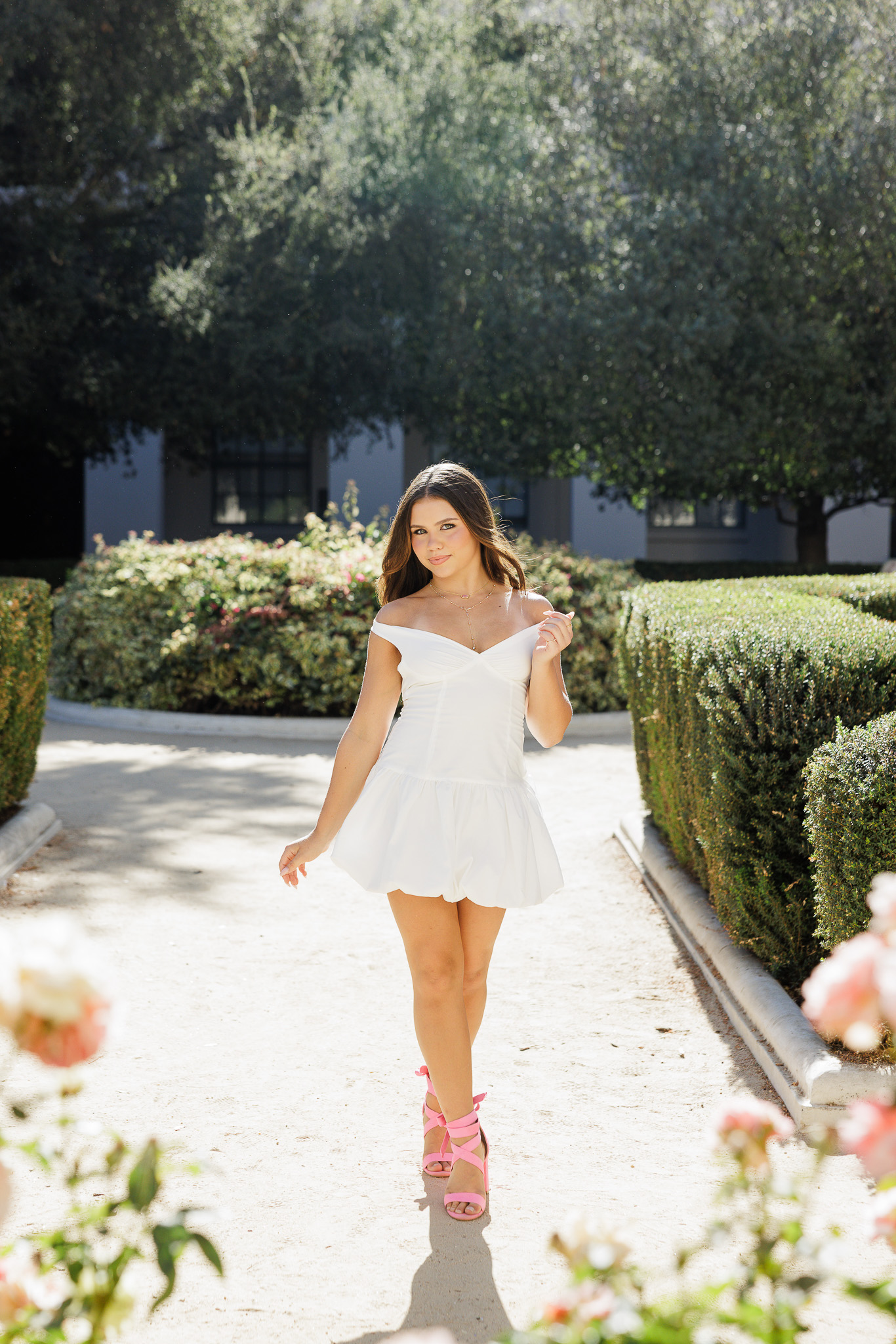 teen girl walking toward the camera in a white mini dress and pink heels between hedgerows with roses in the foreground during her Pasadena California senior pictures