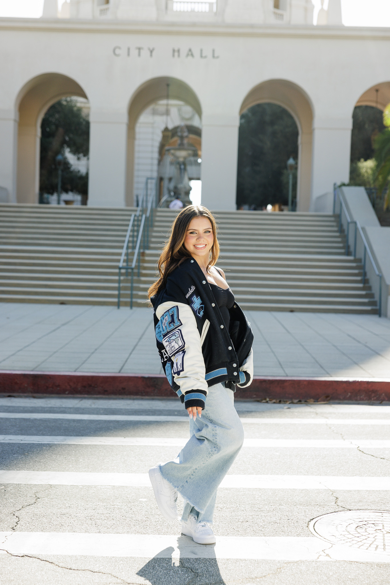 teen girl in her Saugus High School Class of 2027 letterman jacket, light jeans, and white sneakers in front of Pasadena City Hall arches and staircase
