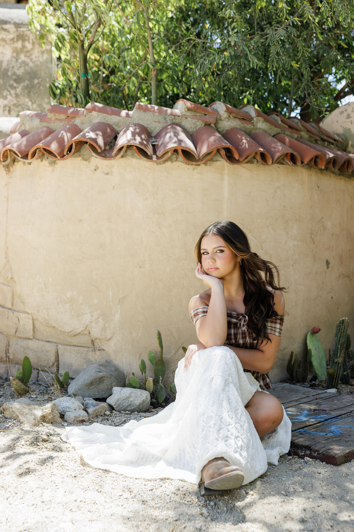 teen girl crouching in her plaid top and white skirt next to a curved stucco wall with terra cotta tile and cacti during her Santa Clarita senior portrait session