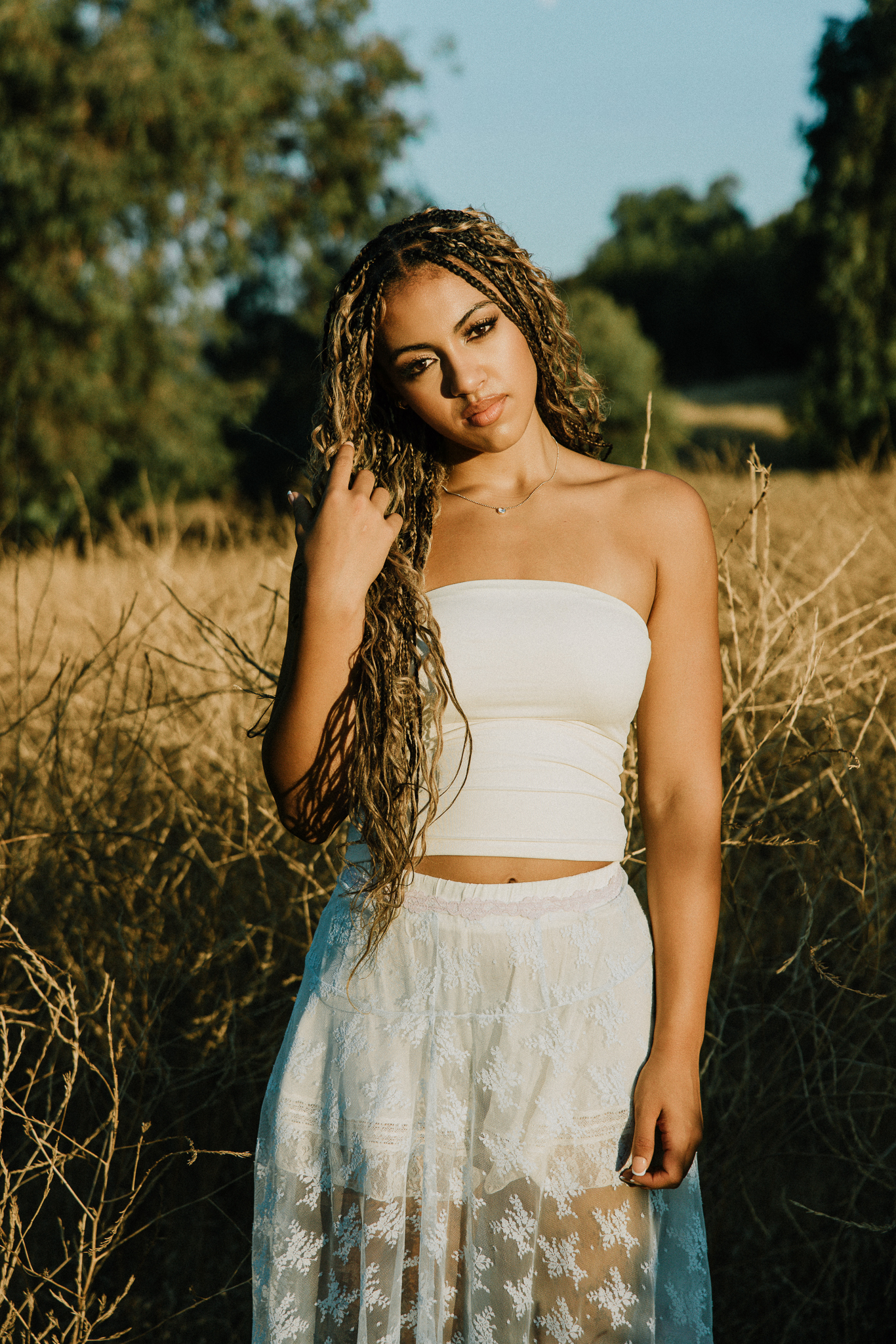 Senior with long braids standing in a golden field of tall dry grass in a white tube top and white lace maxi skirt at golden hour