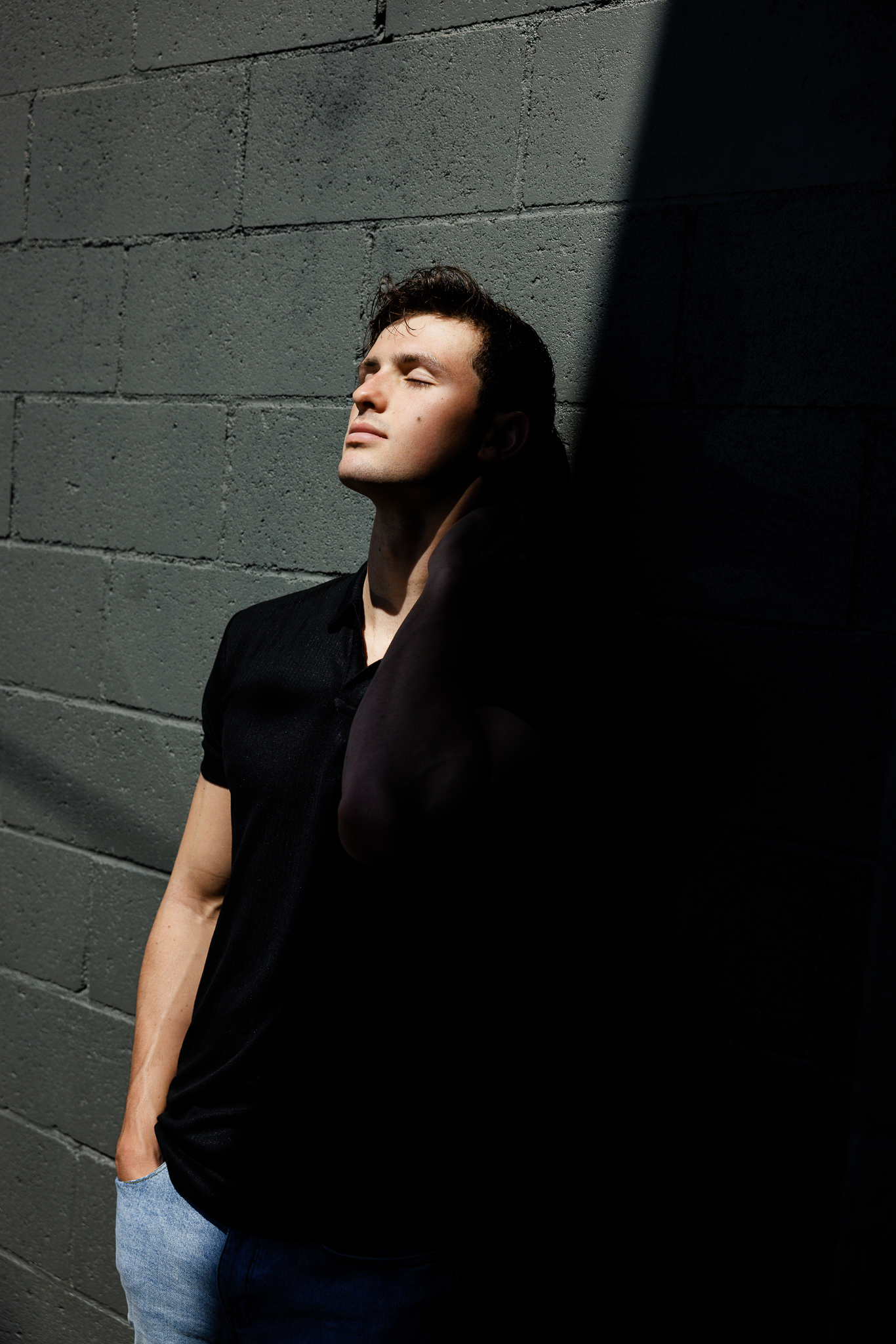Senior guy in a black polo leaning against a gray block wall in hard afternoon shadow
