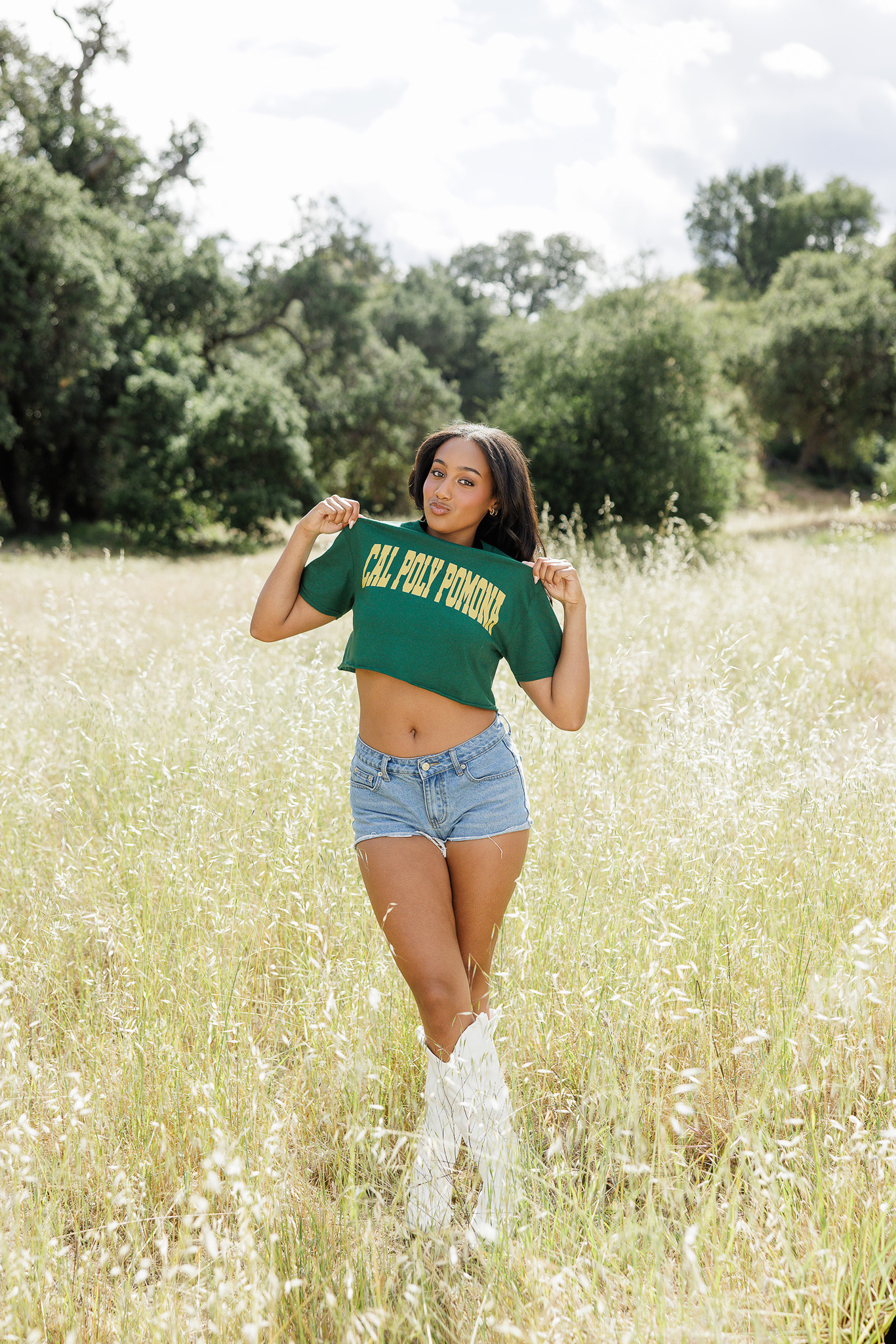 Senior girl holding out her green Cal Poly Pomona shirt in a grassy field in white cowboy boots