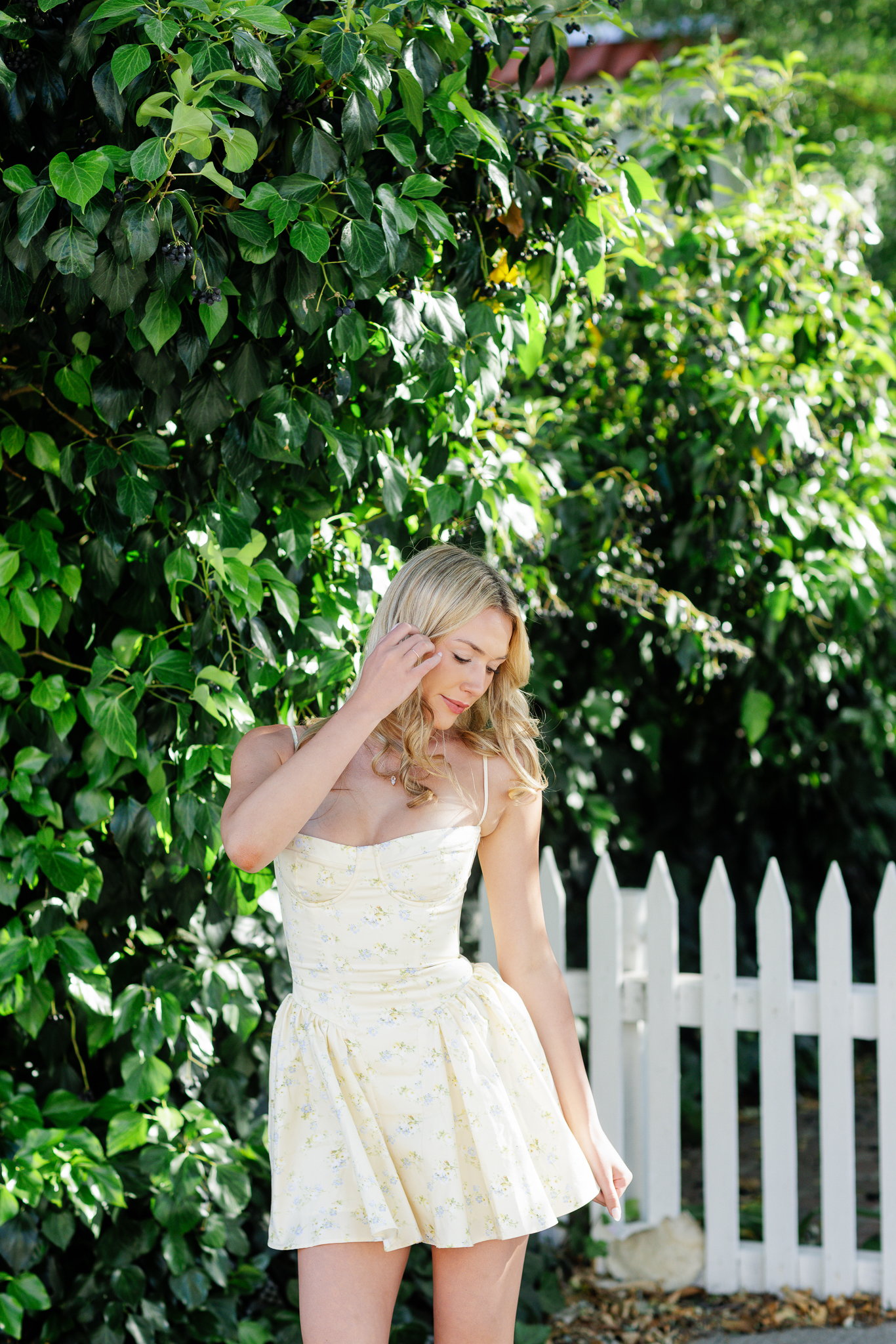 Blonde senior in a yellow floral corset mini dress standing beside a white picket fence and green berry vine with soft backlight