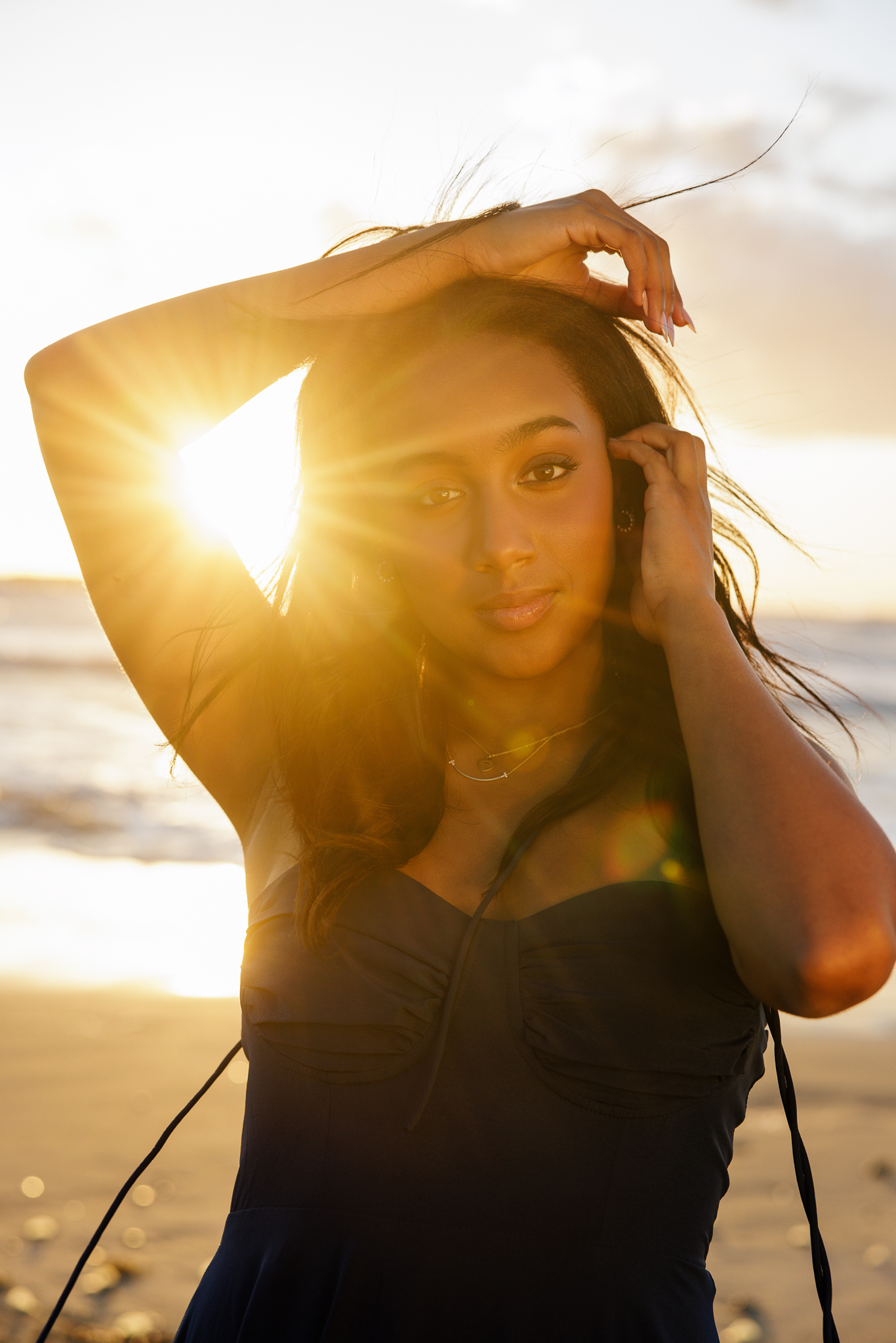Senior with dark hair in a dark blue dress posing on the beach at sunset with golden sun flare bursting behind her