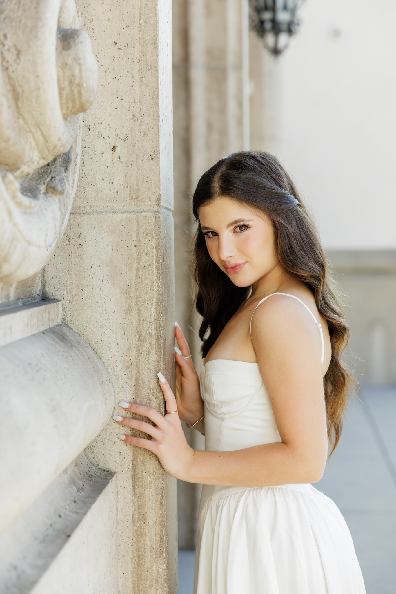 Close-up senior portrait of a brunette girl in a white dress leaning against an ornate stone column and looking directly at the camera during her senior photo session in Southern California